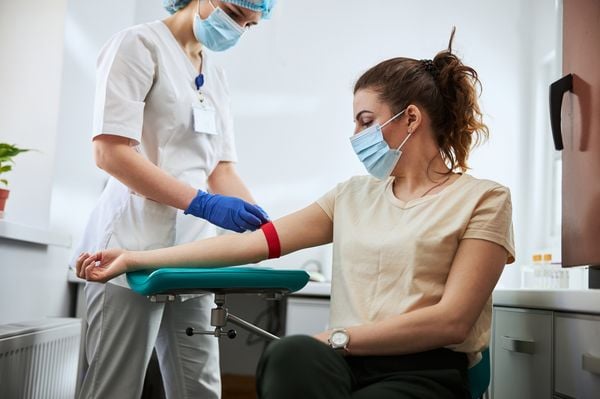 Mulher branca veste camiseta clara e está sentada esperando para fazer exame de sangue, enquanto enfermeira, vestida de branco e com pele branca, amarra um elástico no braço da paciente